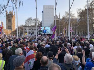 Nigel Farage taler foran Parlamentet i London. Ses på storskjerm foran mange mennesker. Foto