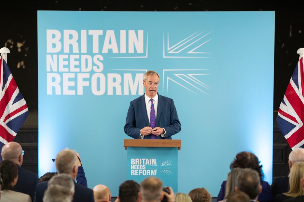 Bilde av Nigel Farage på talestolen med plakat for Reform UK bak seg.