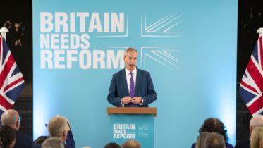 Bilde av Nigel Farage på talestolen med plakat for Reform UK bak seg.